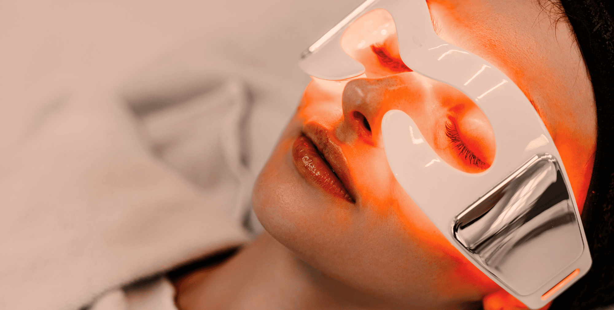 Flat-lay of an LED red light therapy mask, serum bottle and headphones on a linen surface