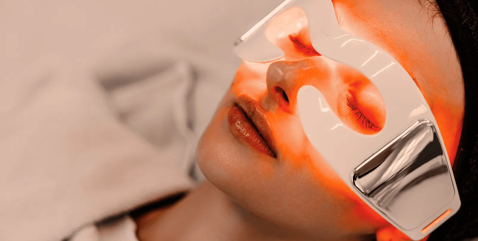 Flat-lay of an LED red light therapy mask, serum bottle and headphones on a linen surface